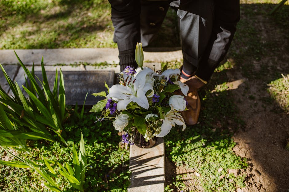 A person dressed in dark clothing, including a black jacket and grey trousers, is seen from above as they place a floral arrangement at a gravesite in West Norwood Cemetery. The bouquet features large