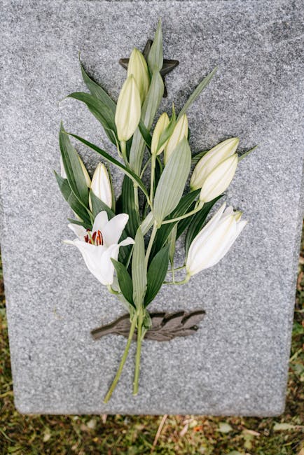 A bouquet of fresh white lilies with prominent red stamens, arranged with long, slender green leaves and closed lily buds, laid flat against a gray stone surface, likely for a floral gift or flower de
