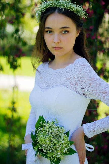 A young woman in a white lace wedding dress holds a small bouquet of lily of the valley with dark green leaves, standing outdoors in a garden setting with lush greenery and pink flowers in the backgro